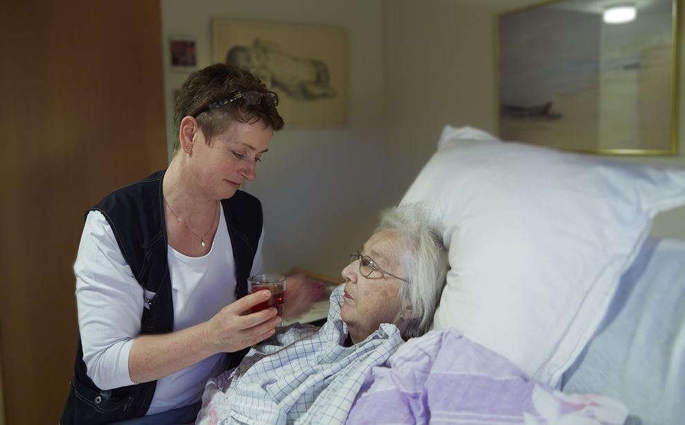 Kvindelig social- og sundhedsassistent giver juice til liggende ældre kvinde